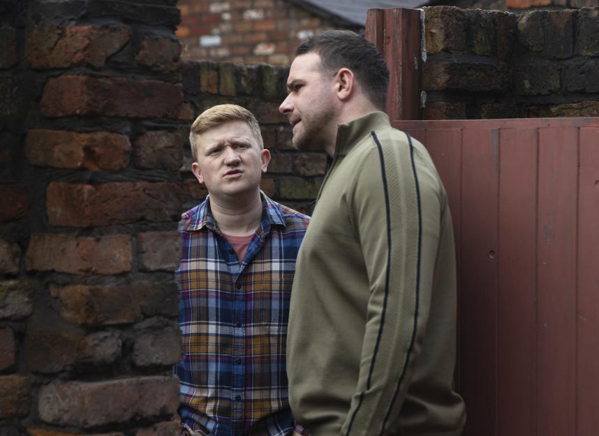 Two men talking by a brick wall.