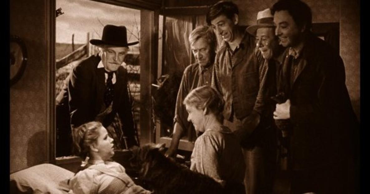 Scene from a film depicting a group of men surrounding a young girl lying in bed. A man in a hat stands behind her.