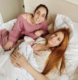 A woman breastfeeding a newborn baby while another woman sits beside them on a bed.