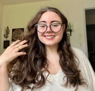 Woman with long brown hair, wearing glasses and a cream-colored sweater, touching her hair.