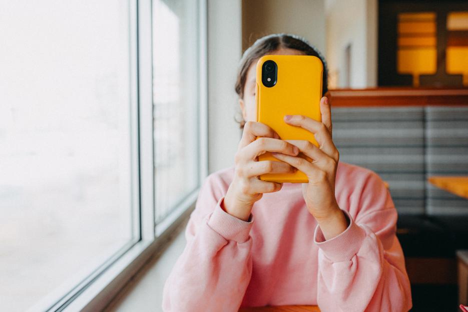 Girl sitting at a table by a window using a yellow smartphone.