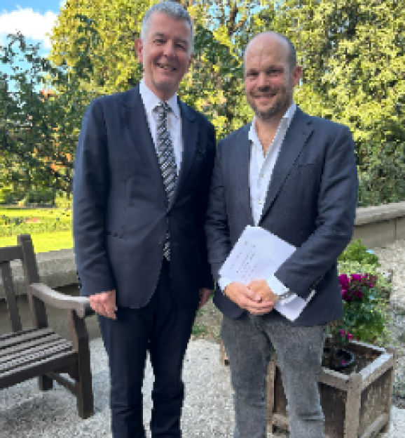 Jermoe in a suit and tie, smiling next to a man in a dark jacket holding papers.