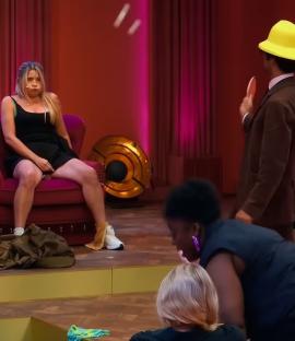 Woman sitting on a chair on a stage, a man in a yellow hat holding a ping pong paddle, and other people watching.