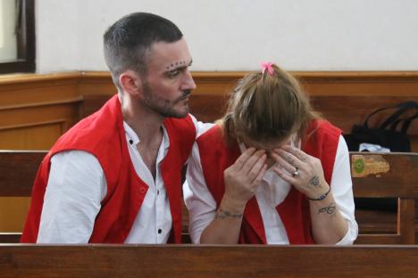 A man and woman in red vests sit in court; the woman is crying.