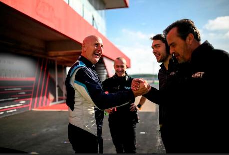 Two men shaking hands at a racetrack, with another man in the background.