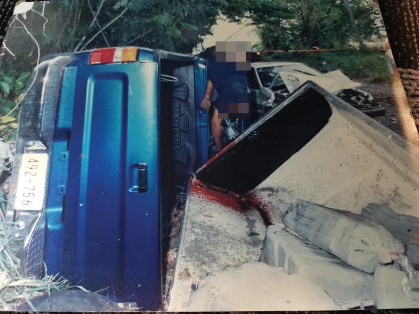 Evidence from the Cartel: A blue truck on its side, a body hanging from the door, a pool of blood on the ground, and several white packages, possibly drugs, next to a concrete barrier.