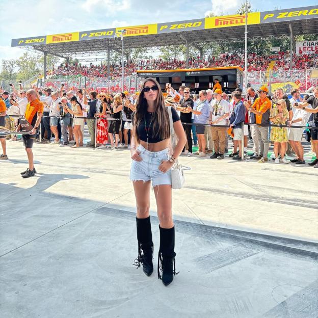 A woman in sunglasses, a black crop top, denim shorts, and black boots stands on concrete in front of a crowded grandstand with "P ZERO PIRELLI" banners.