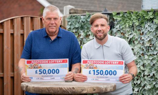 Two men holding oversized checks from the Postcode Lottery; one for £50,000 and the other for £150,000.