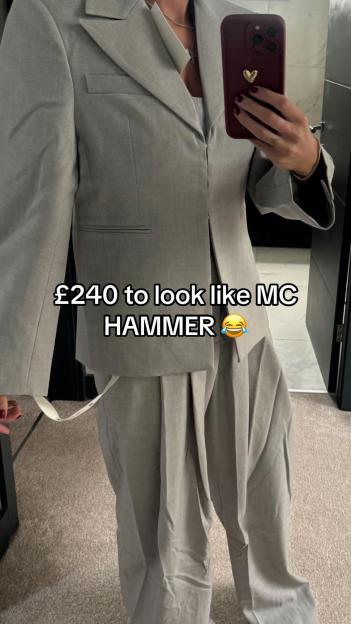 A person in an oversized gray pantsuit holds a phone to take a selfie.