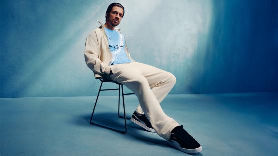 Man sitting on chair wearing cream pants, light blue shirt, and black shoes.