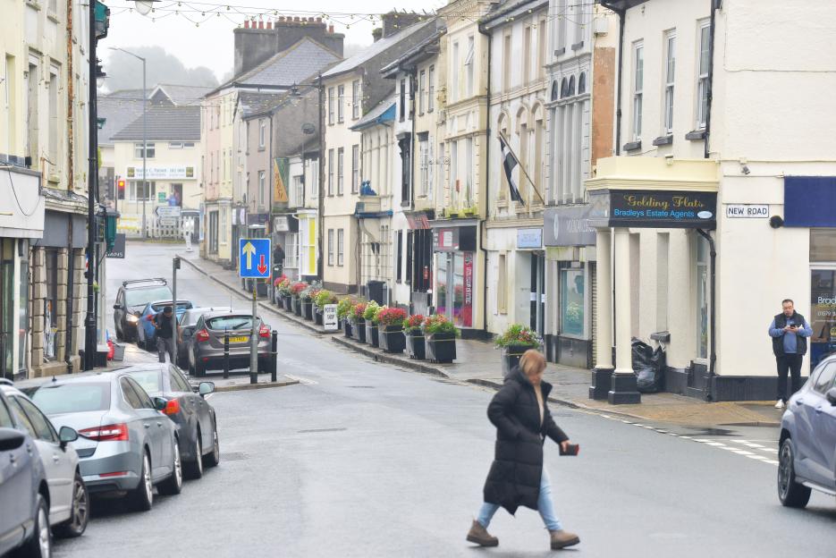 Street in Callington, Cornwall, lined with buildings and parked cars, with a woman walking in the foreground.