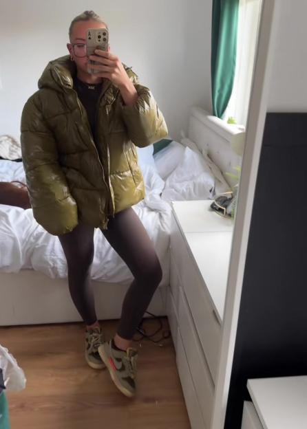 Woman modeling an olive green puffer jacket in a bedroom.
