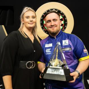 A man and woman stand together; the man holds a Premier League darts trophy.