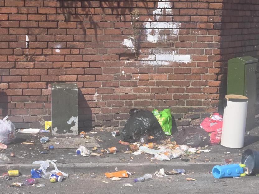 Fly-tipped waste, including trash bags and litter, on a street against a brick wall.