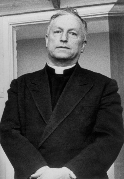 Black and white photo of Father Anthony Joseph Crean wearing a clerical collar and jacket, standing with hands clasped.
