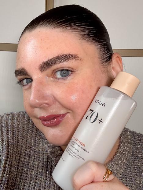 A woman with dewy skin, dark hair slicked back, and wearing a glittery top, holds a bottle of Anua 70+ Rice 70 Glow Milky Toner.