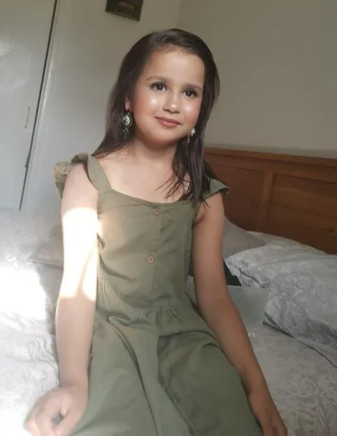 A young girl with long dark hair, wearing a green dress and earrings, sits on a bed and smiles.