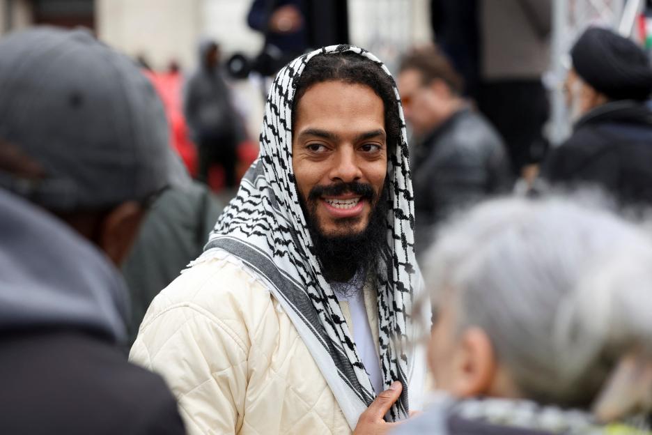 Bobby Vylan smiling and wearing a keffiyeh and a quilted jacket, at a protest in London.