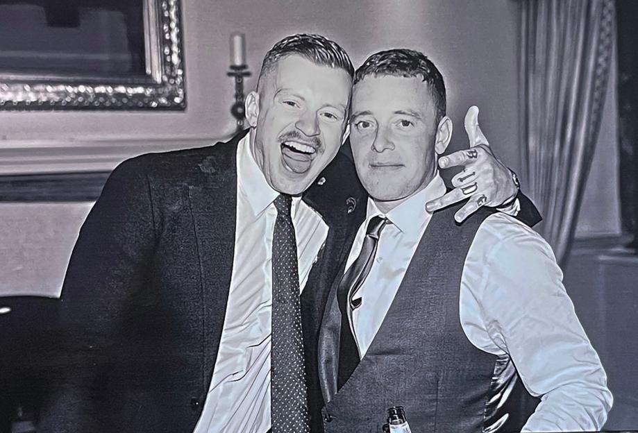 Black and white photo of Olympic swimmer Adam Peaty and his brother James embracing, Adam on the left laughing, and James on the right with a "BJD" tattoo on his hand.