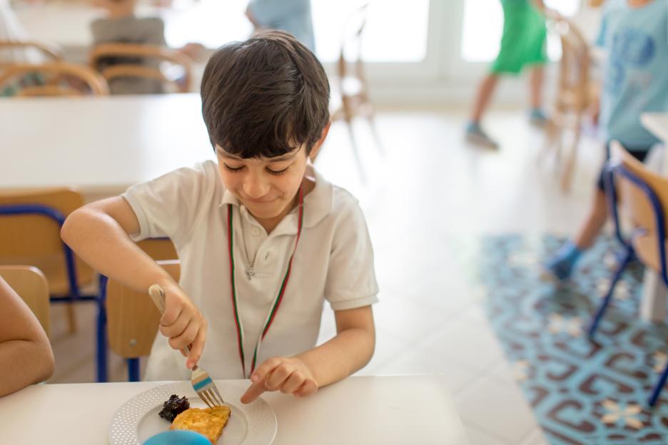Boy sitting at a table with a plate of breakfast food, using a fork to eat.