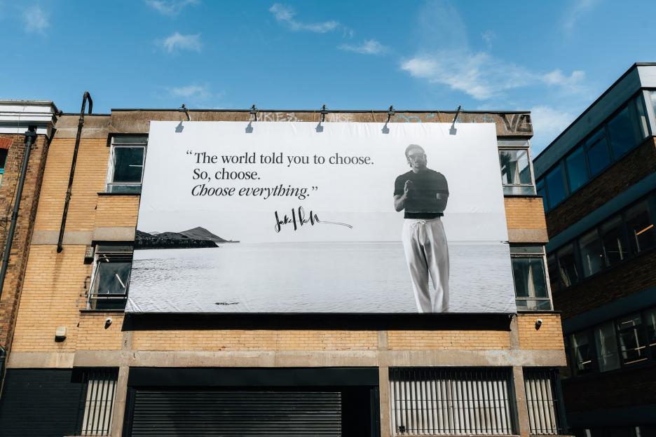 A large billboard on a brick building features a black and white image of a person standing by a body of water and the quote: "The world told you to choose. So, choose. Choose everything."