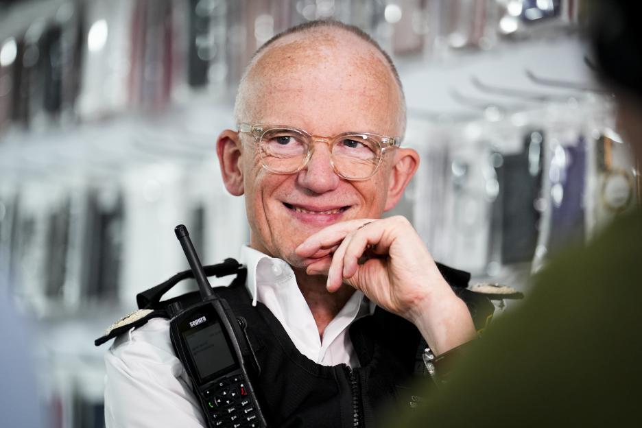 Metropolitan Police Commissioner Sir Mark Rowley, wearing glasses and a uniform, smiles and looks to his right, with a radio attached to his chest.
