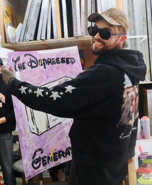 A man with a beard, sunglasses, and a cap smiles while holding up a purple painting that reads "THE DEPRESSED" and "GENERA 2025" in a workshop.