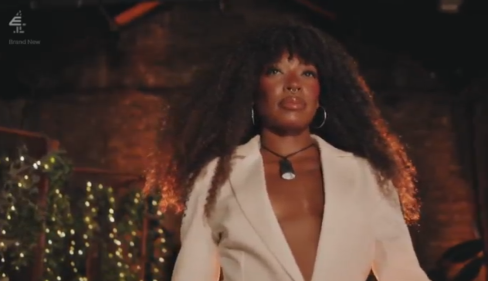 A Black woman with long, curly dark hair and a septum piercing, wearing a white blazer and a dark necklace, looks forward.