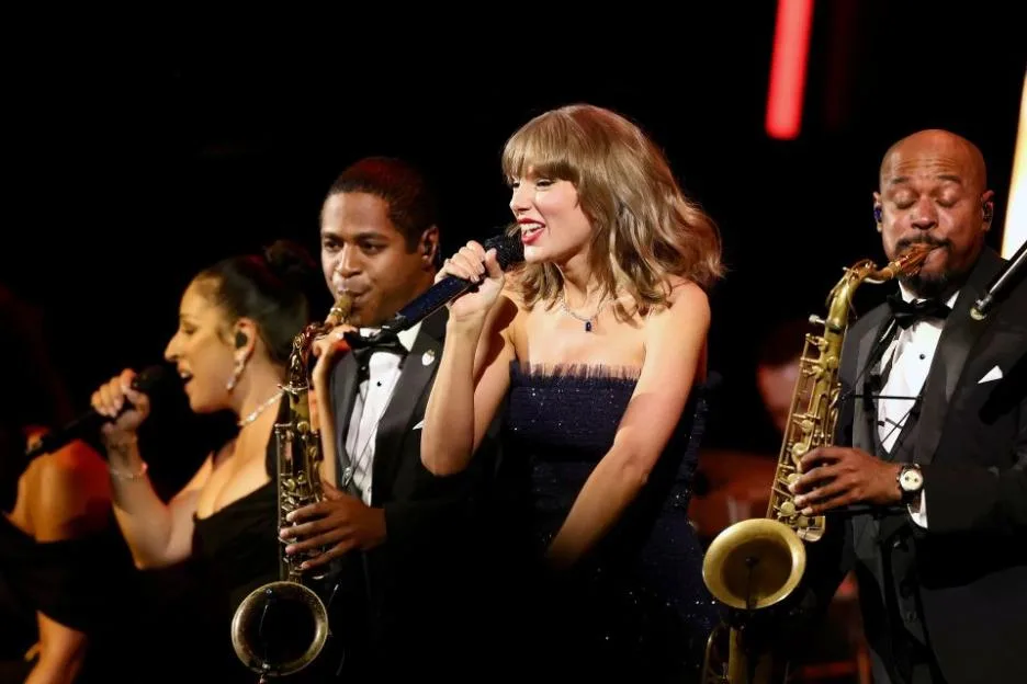 Taylor Swift performs onstage, singing into a microphone, accompanied by musicians playing saxophones.