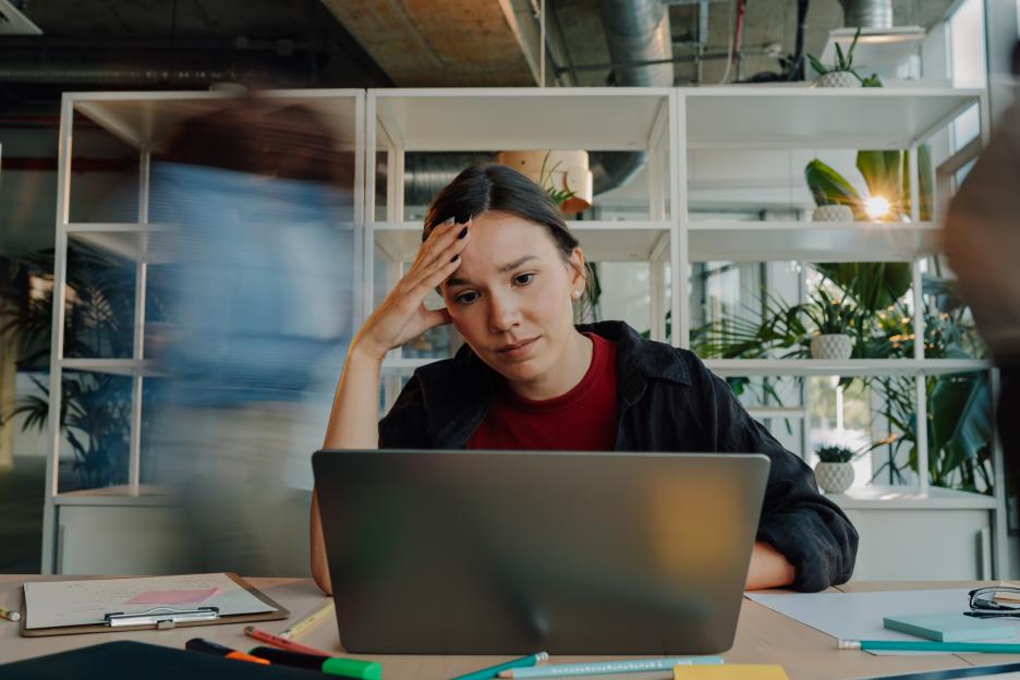 Stressed young woman working on laptop in busy office