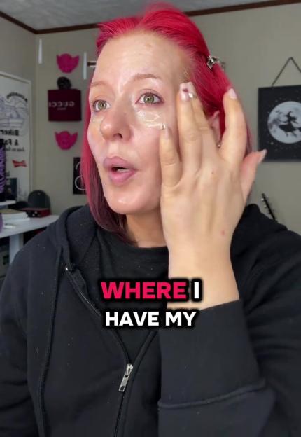 A woman with bright pink hair applies a clear product to her face.