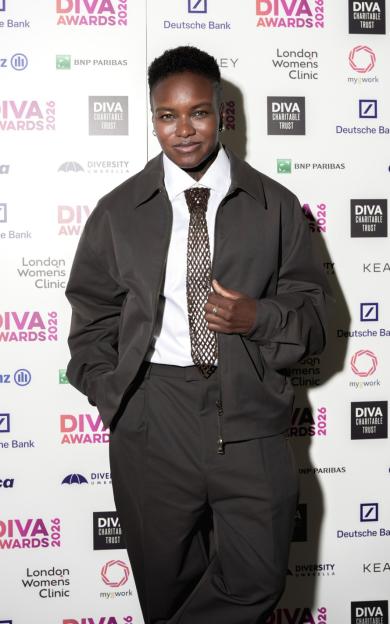 Kae Tempest posing in a dark brown suit and a beaded tie in front of a white step-and-repeat banner with various logos.