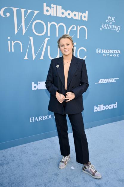 Jojo Siwa in a dark suit with no shirt underneath, standing on a light blue carpet in front of a Billboard Women in Music sign.