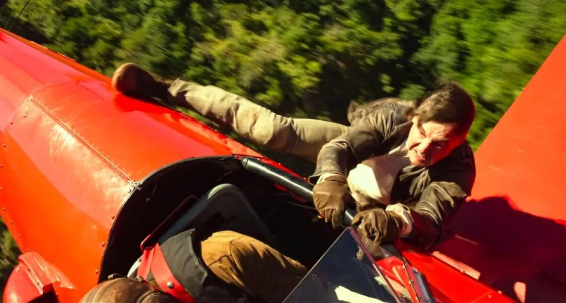 Tom Cruise hangs onto the outside of the cockpit of a flying red airplane in a scene from Mission Impossible - The Final Reckoning.