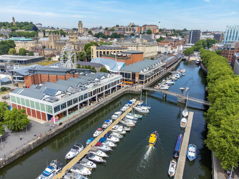 Aerial view of the Bristol Harbour area, with boats docked along the river, buildings, and a Ferris wheel in the background.