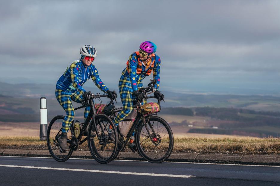 Two cyclists in matching tartan outfits ride bicycles on a road with a vast landscape in the background.