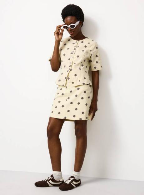 A model wears a short-sleeved, collared button-up top and matching mini skirt, both light yellow with dark polka dots. She also wears white cat-eye sunglasses and brown sneakers.