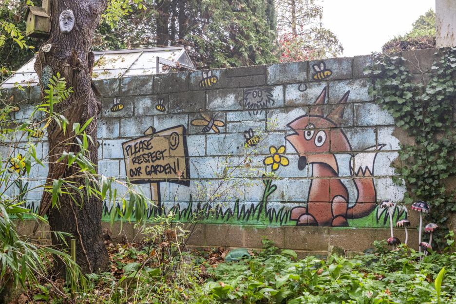 Illustration of a fox, bees, and a sign that reads "Please Respect Our Garden" painted on a brick wall next to a tree and ivy.