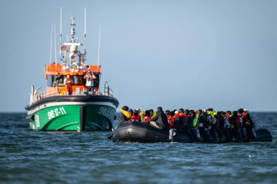 A rescue boat approaches a crowded inflatable dinghy carrying migrants in life jackets at sea.