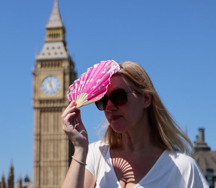 A woman uses a pink fan to shade her face from the sun, with the Big Ben clock tower blurred in the background.