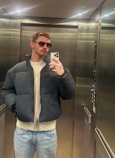 Jack Keating taking a mirror selfie in an elevator, wearing a puffer jacket and sunglasses, with text about returning home and traveling to the US.