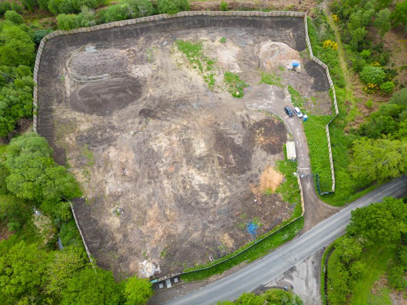 Aerial view of the demolished Four Houses Corner Caravan Site, a large dirt lot surrounded by a fence and green trees, with a road running alongside it.
