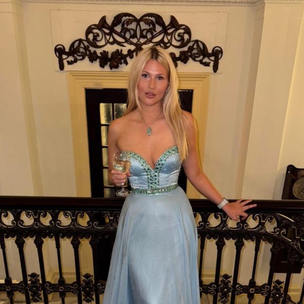 A woman in a light blue strapless dress with a jeweled bodice holds a wine glass while standing by a dark ornate railing.