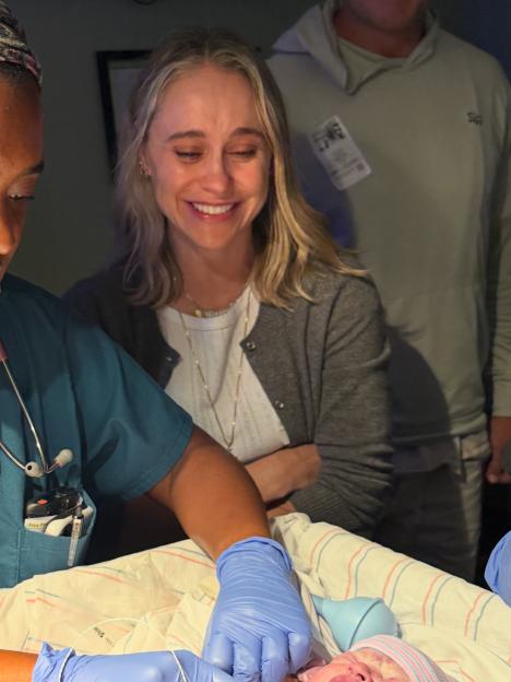 A woman in scrubs, a smiling woman in a gray cardigan, and a man in a gray hoodie look down at a newborn baby.