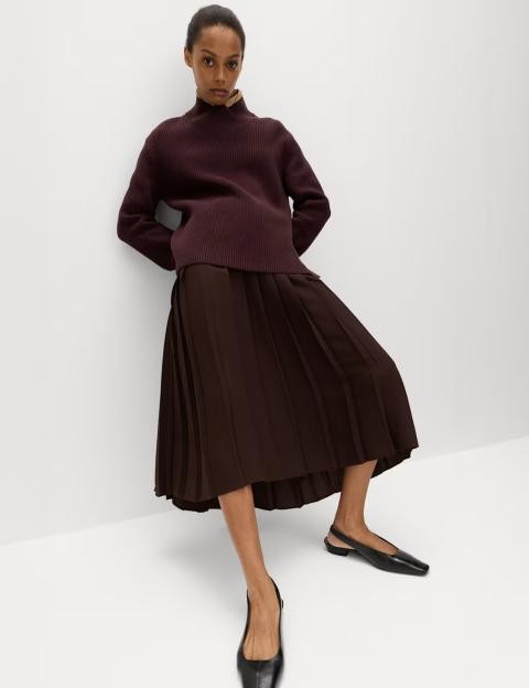 A woman wearing a dark red ribbed longline jumper and a dark brown pleated skirt with black flat shoes.
