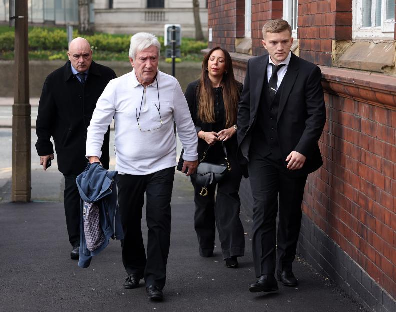 Four people, two men, one man in a white polo shirt, another man in a black suit, and a woman in black clothes, walk past a brick wall.