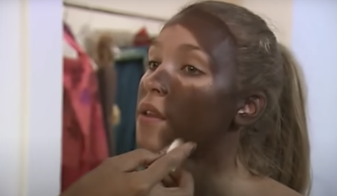 A woman having dark makeup applied to her face, leaving some of her natural skin tone visible.