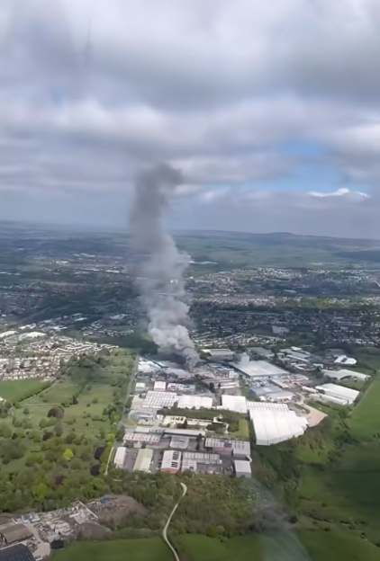 Aerial view of a large fire with dark smoke rising from a factory in an industrial area, surrounded by green fields and residential buildings under a cloudy sky.