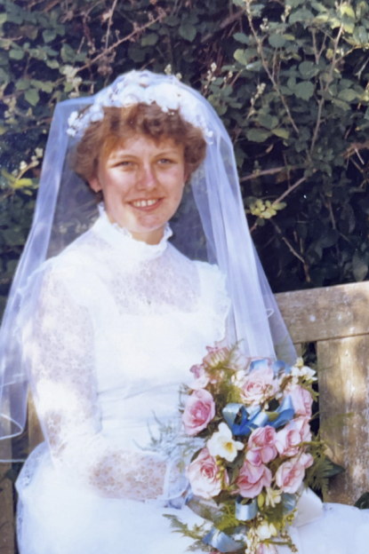 Joy Boulton on her wedding day, wearing a white dress and veil, holding a bouquet of pink and blue flowers, smiling at the camera.