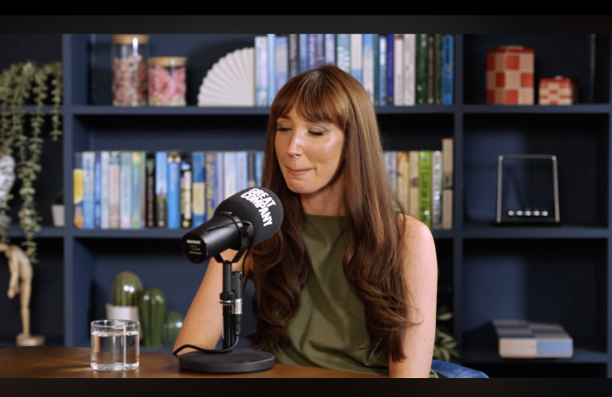 Megan McKenna sitting at a table with a microphone in front of a bookshelf, with her eyes closed, as if in tears.